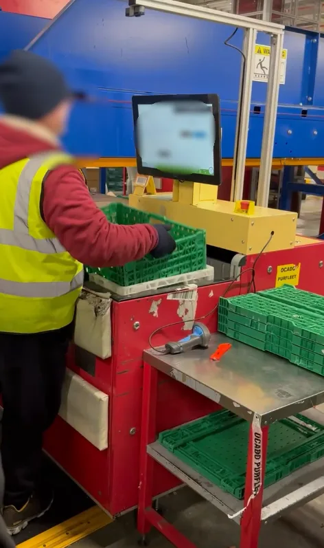 A Decant operative working at the automation station, reading the on-screen prompt beside the conveyor.
